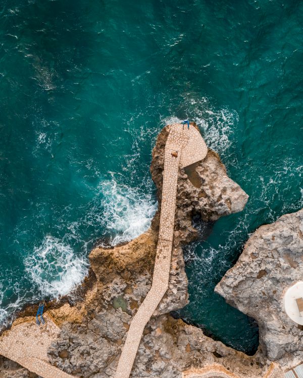 Aerial view of rugged rocky coastline with a concrete jetty extending into turquoise water, small white circular platforms at the edge, waves crashing below.