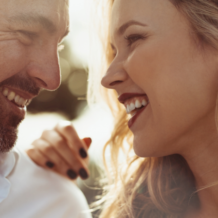 Two adults smiling warmly at each other, close up, suggesting affection or a conversation, with sunlight behind them and a relaxed vibe.