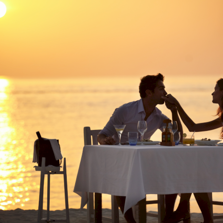 A romantic sunset beach dinner for two, candlelit table by the sea as the couple share a kiss at sunset.