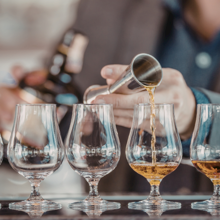A bartender pours whiskey into a row of tulip glasses on a bar counter, with a line of amber liquid forming as he tilts the bottle.