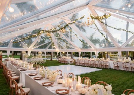 A modern outdoor wedding venue with white chairs, wooden arbors, string lights, and greenery under a clear glass canopy.