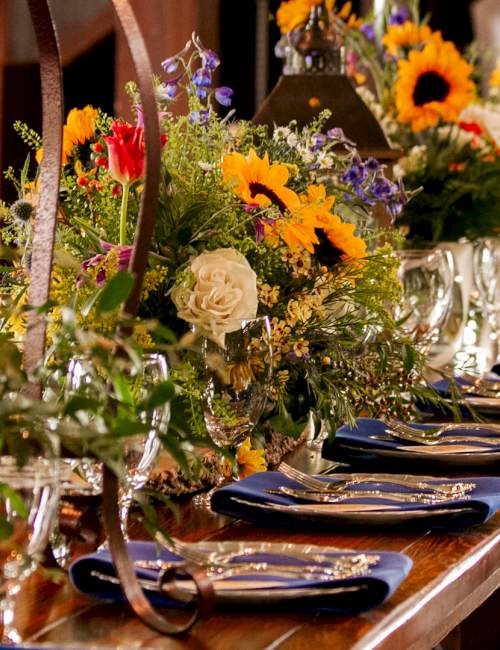 A long dining table set for a formal feast with colorful flower centerpieces, folded napkins, and glassware&mdash;elegant and festive.