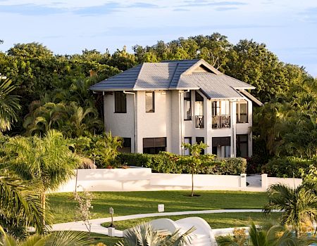 A modern two-story beige house with a gray roof, surrounded by palm trees and a white fence in a lush tropical setting.