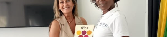 Two women stand indoors, one handing the other a small book labeled &ldquo;Wine&rdquo; with a colorful grape graphic on the cover, both smiling.