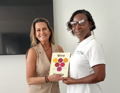 Two women stand indoors, one handing the other a small book labeled &ldquo;Wine&rdquo; with a colorful grape graphic on the cover, both smiling.