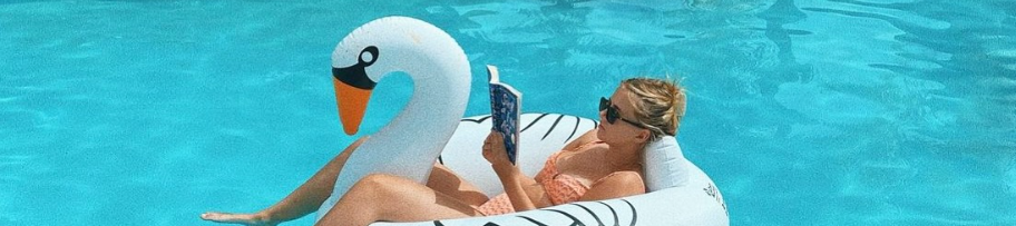 A person floats on a swan-shaped inflatable in a bright pool, reading a book with sunglasses on, summer vibes all around.