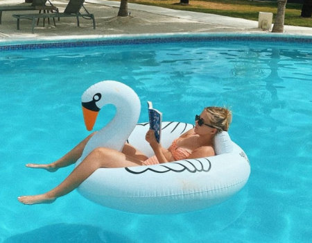 A person floats on a swan-shaped inflatable in a bright pool, reading a book with sunglasses on, summer vibes all around.