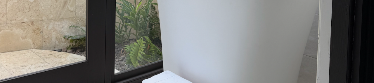 A modern bathroom with a freestanding white bathtub, a small wooden stool with a folded towel, and a glass door revealing plants outside.