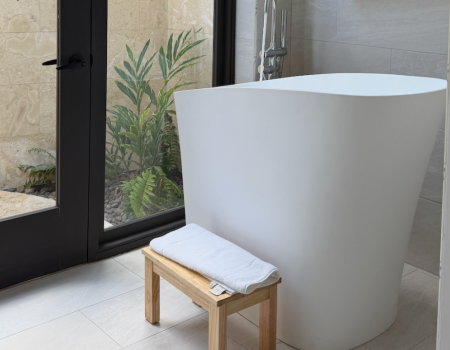 A modern bathroom with a freestanding white bathtub, a small wooden stool with a folded towel, and a glass door revealing plants outside.