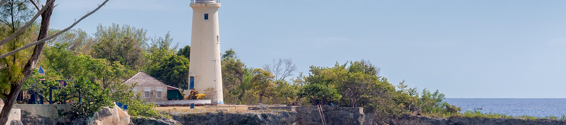 A scenic coastal scene with a white lighthouse on rocky cliffs, turquoise water, leafy trees, and a clear blue sky, evoking calm seaside vibes.
