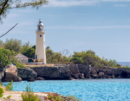 A scenic coastal scene with a white lighthouse on rocky cliffs, turquoise water, leafy trees, and a clear blue sky, evoking calm seaside vibes.