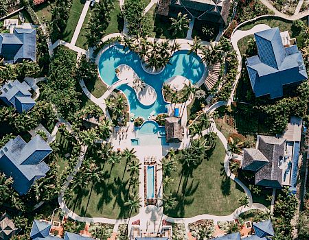 Aerial view of a luxury resort with a large turquoise pool, gardens, palm trees, and surrounding buildings.