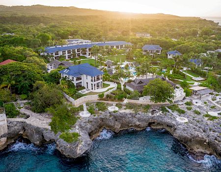 Aerial view of a tropical seaside resort with villas, pools, and a rocky cove at sunset.