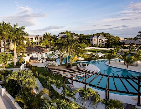 A tropical beachfront resort with palm trees and a large blue swimming pool under a partly cloudy sky.