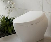 A sleek white modern toilet in a bright tiled bathroom with marble walls and nearby greenery.