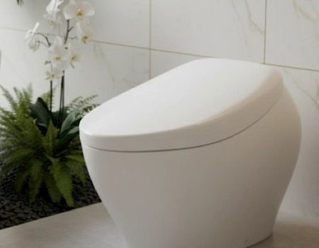A sleek white modern toilet in a bright tiled bathroom with marble walls and nearby greenery.