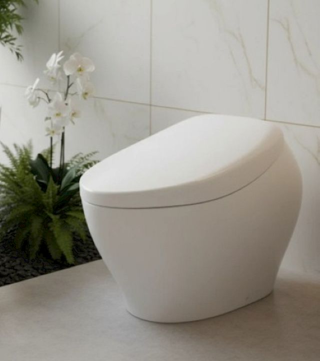A sleek white modern toilet in a bright tiled bathroom with marble walls and nearby greenery.