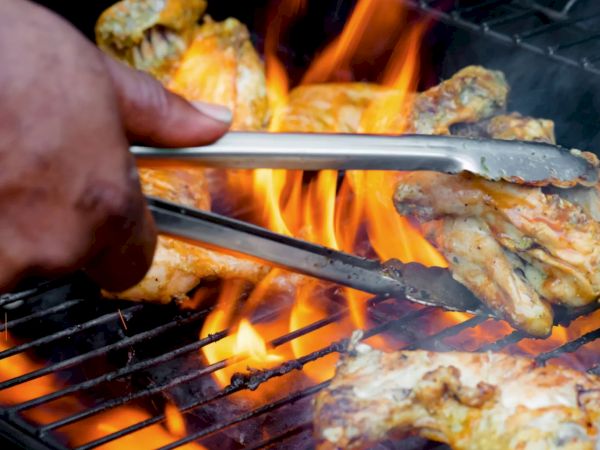 Grilled chicken pieces sizzle over open flames on a charcoal grill, held near the fire by a hand and tongs.