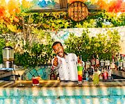A smiling bartender/chef poses poolside behind shelves of colorful liquor bottles and cocktails, with a memorial plaque reading “Only One.”