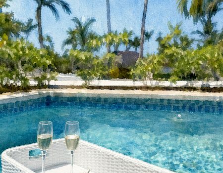 A sunny poolside scene with palm trees, two champagne flutes, and a fruit plate resting on a wicker table.