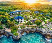 An aerial view of a lush tropical resort with villas, pools, gardens, and a rocky beachfront overlooking sparkling turquoise water.