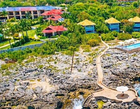 Aerial view of a tropical resort with colorful buildings, pools, and a beach with a wooden walkway winding through rocks and sand.